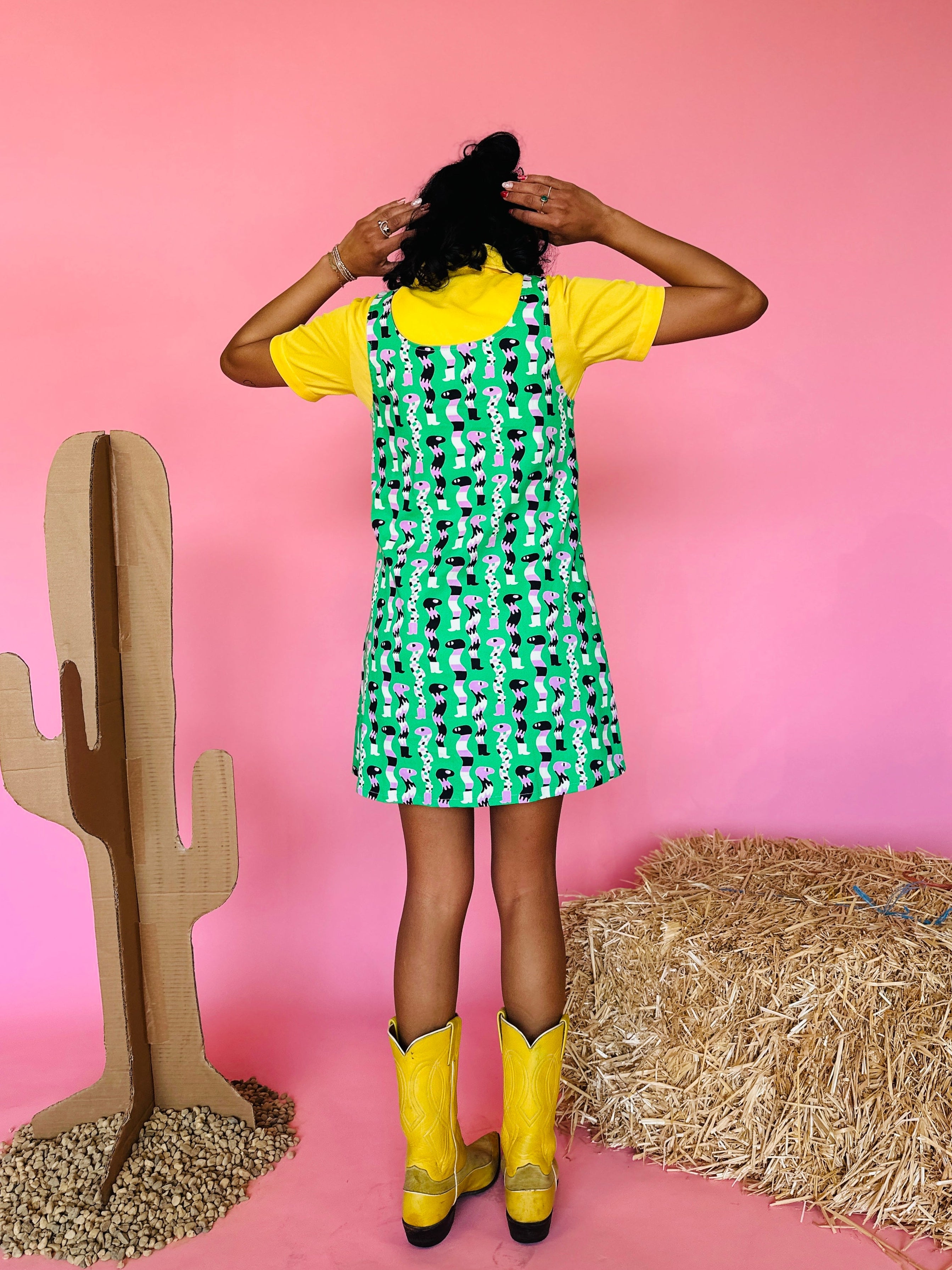 Person wearing a green dress with black patterns and yellow boots standing in front of a pink wall with hay and a cardboard cactus.