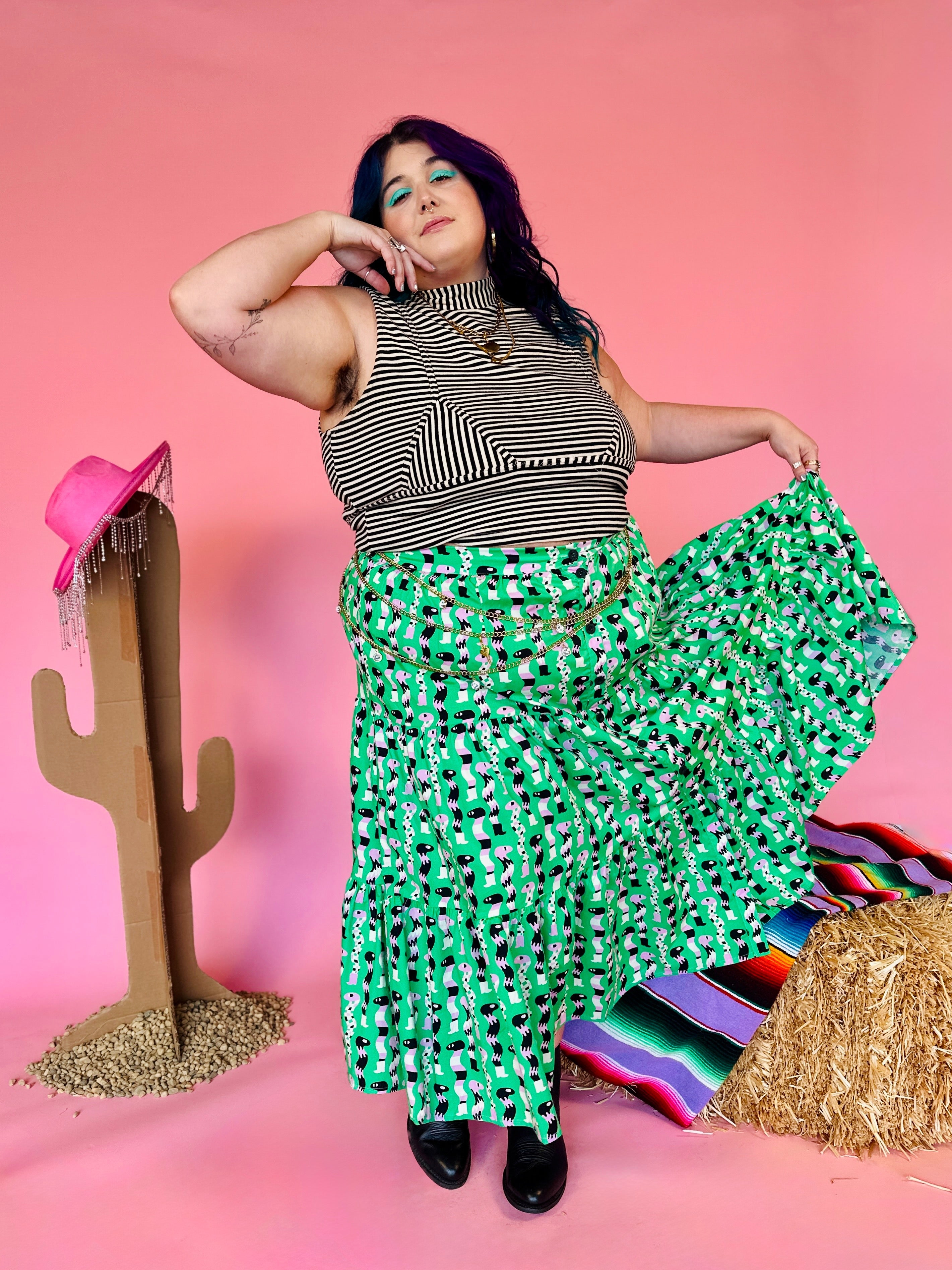 Woman in a green patterned skirt and striped top posing with a cactus and colorful rug on a pink background