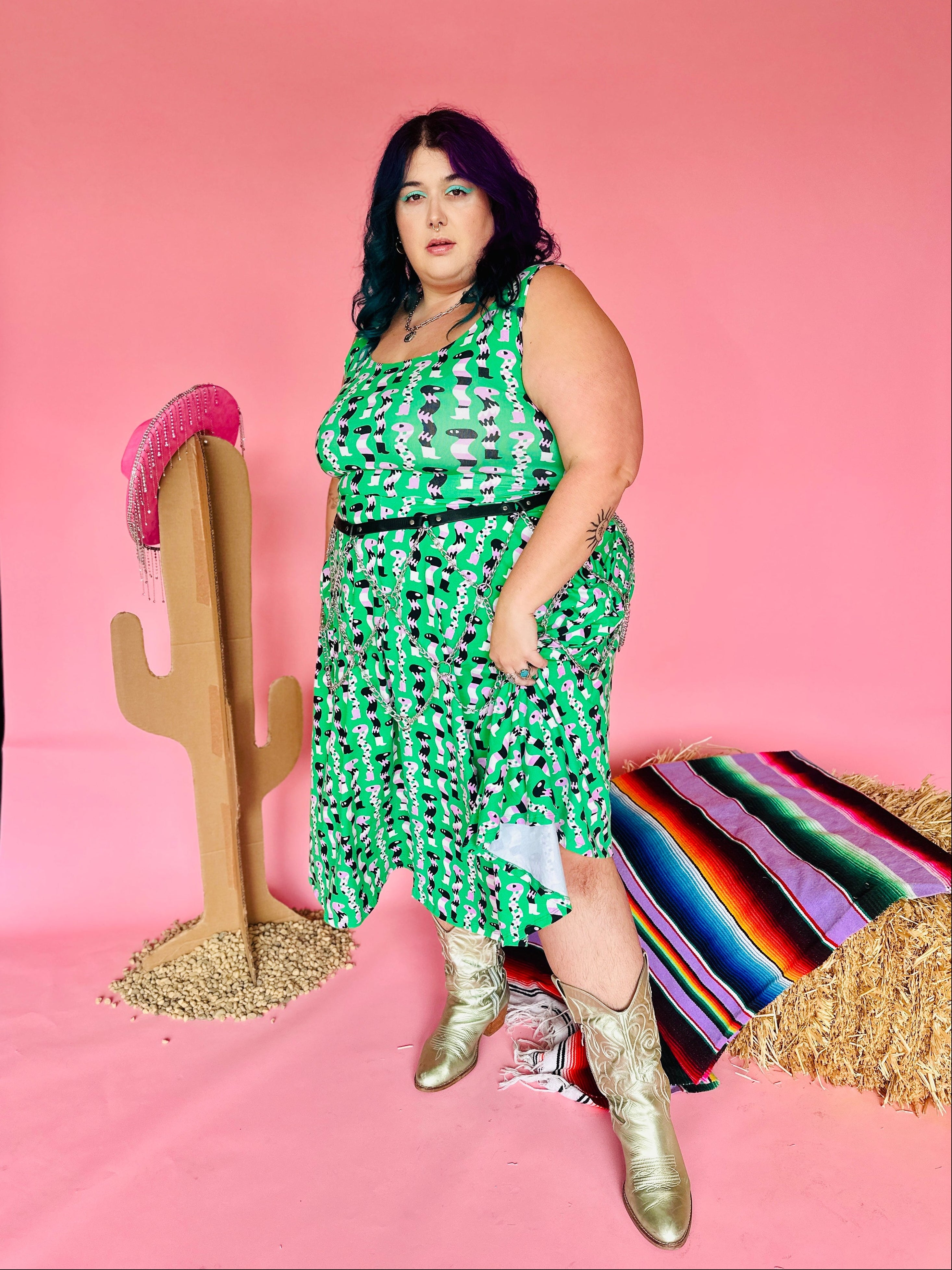 Woman in a green dress standing in front of a pink background with a cactus and serape.