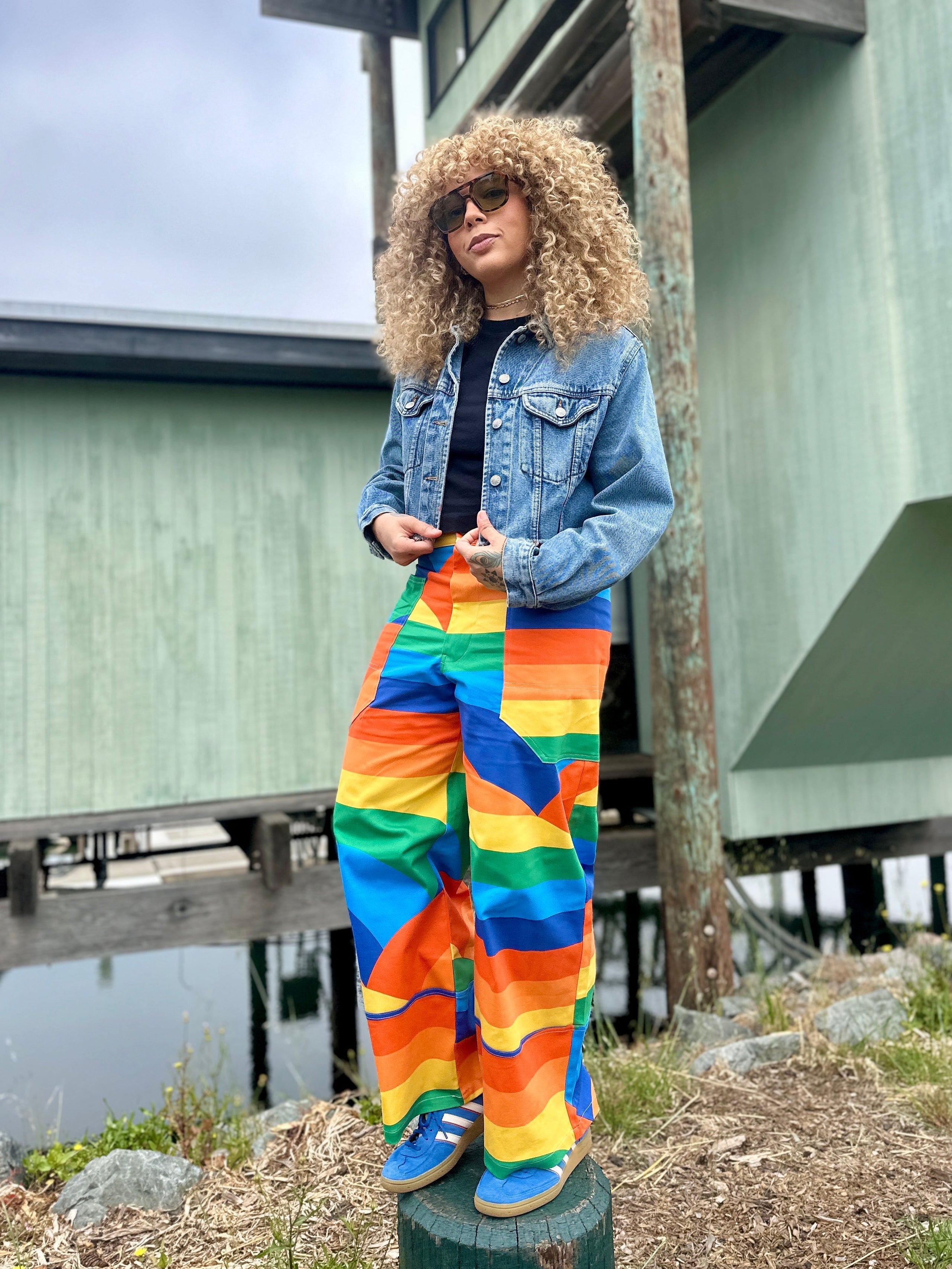 Person wearing a denim jacket and rainbow pants standing outdoors near a building.