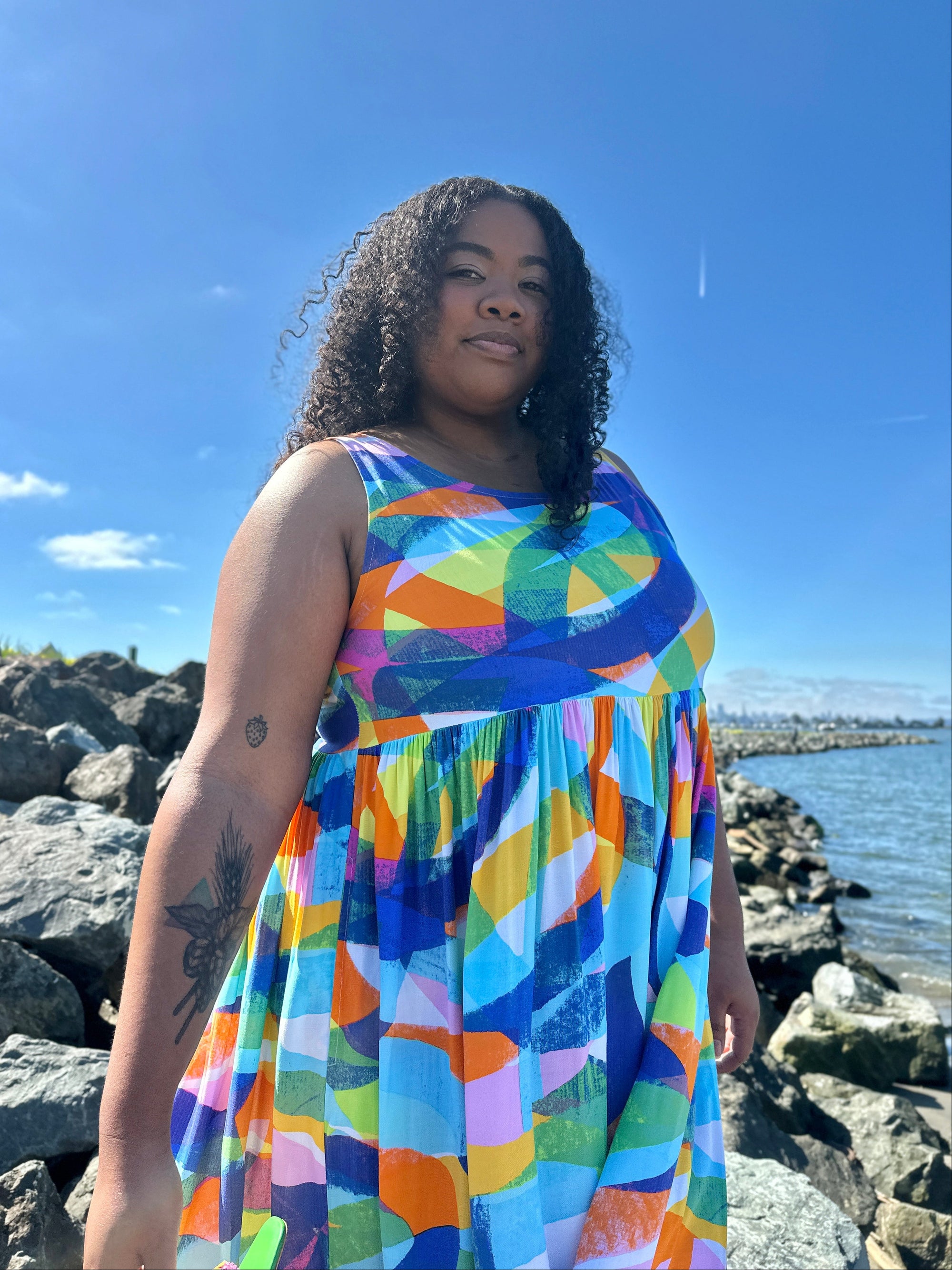 Woman in a colorful dress standing on a rocky shoreline with water and sky in the background