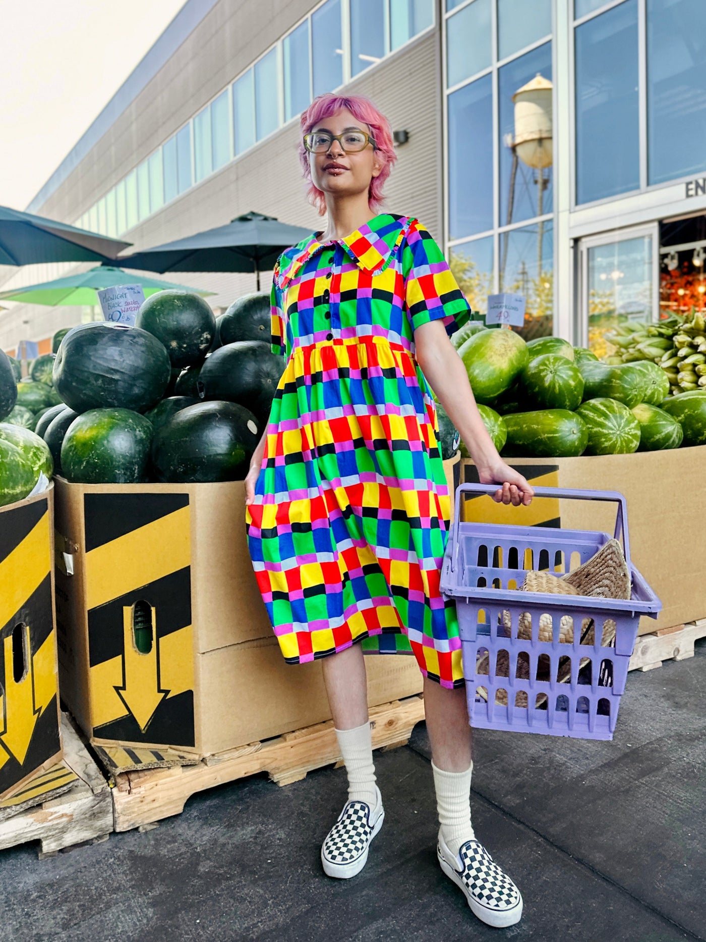 Person wearing a colorful checkered dress standing in front of a produce stand.