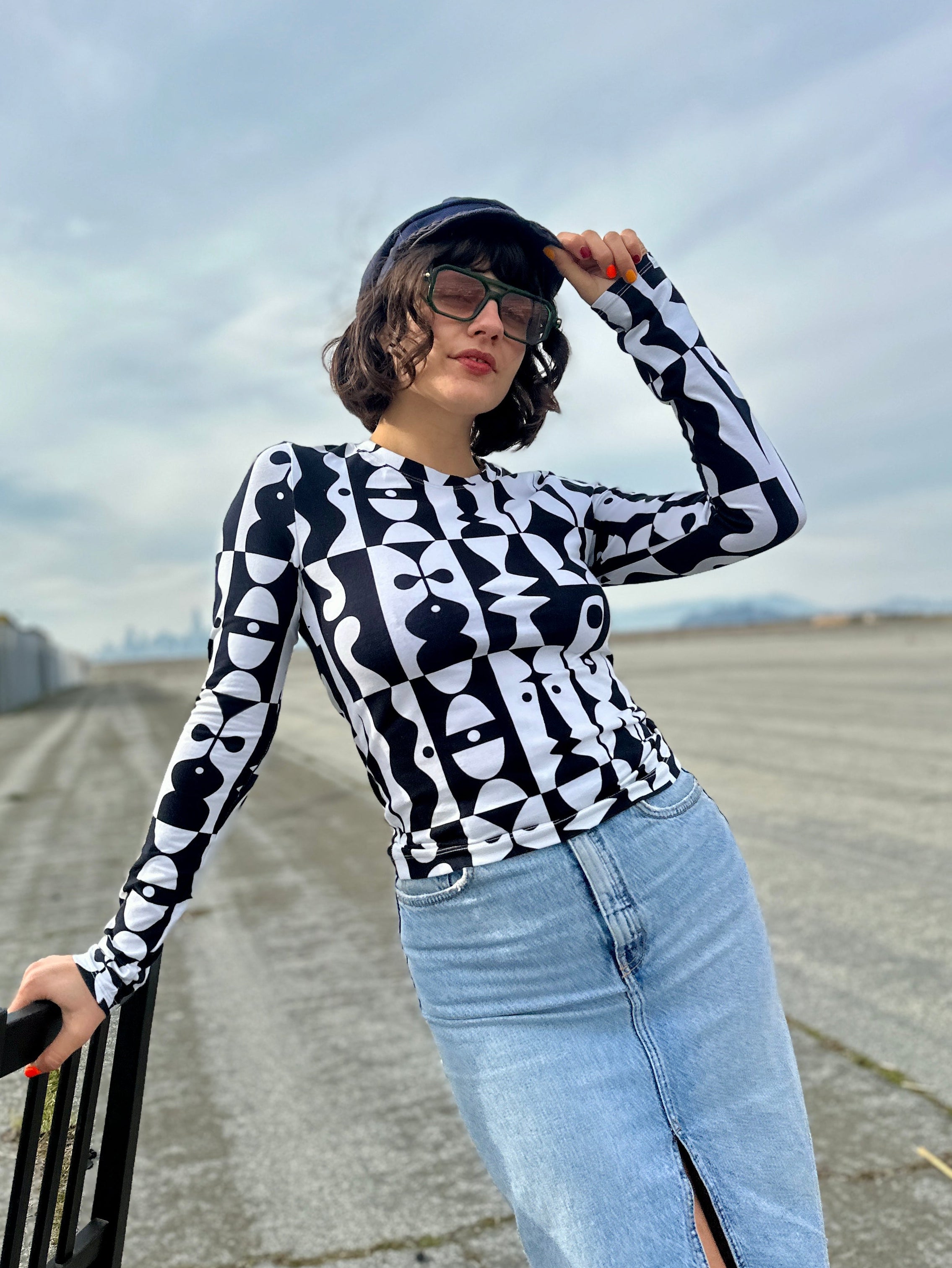 Woman wearing a black and white patterned top and blue jeans standing outdoors with a cloudy sky.
