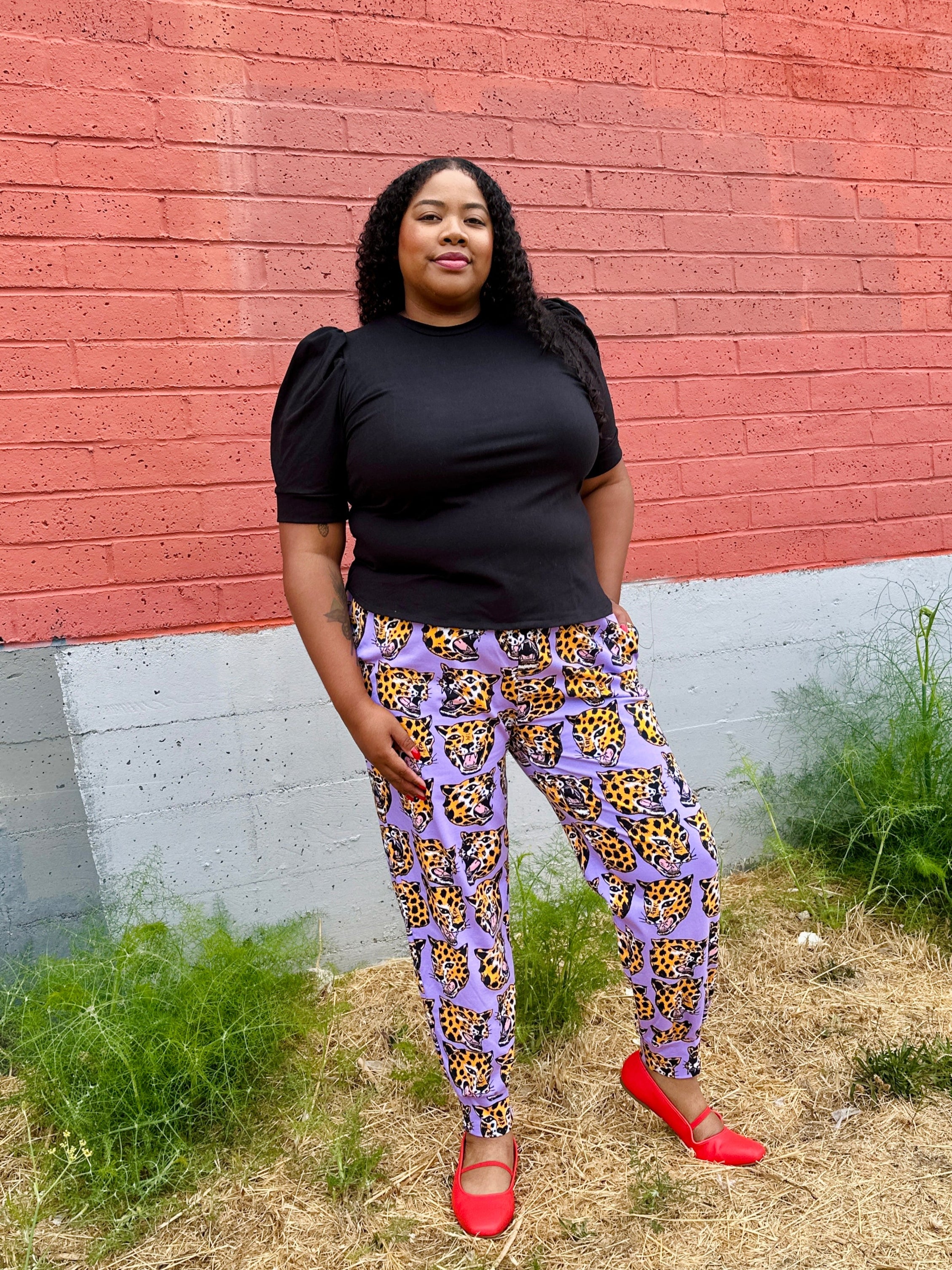Woman wearing a black top and purple patterned pants standing in front of a red brick wall.