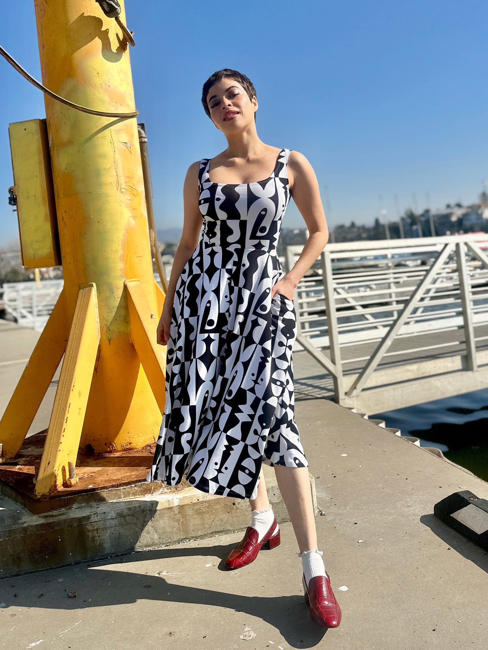 Woman in a black and white patterned dress standing on a dock with a yellow structure and blue sky in the background.