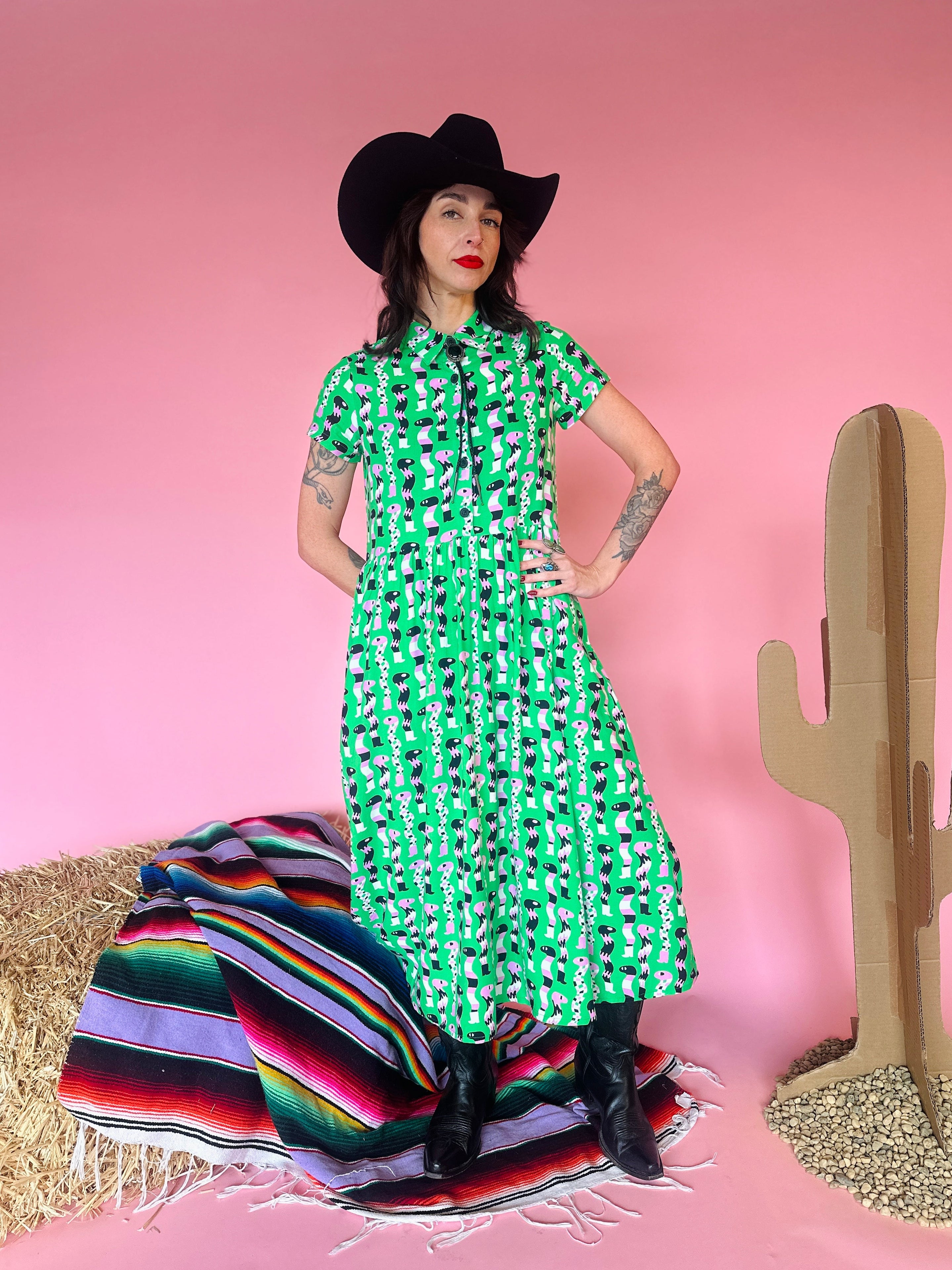 Person wearing a green dress with black patterns, black cowboy hat, and boots in a desert-themed setting with pink background.